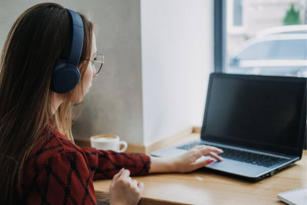 Picture of a girl wearing a headset and navigating her laptop while doing something, together with her coffee in her desk.