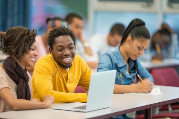 Image of the students looking into the laptop while smiling and writing something in the  paper.