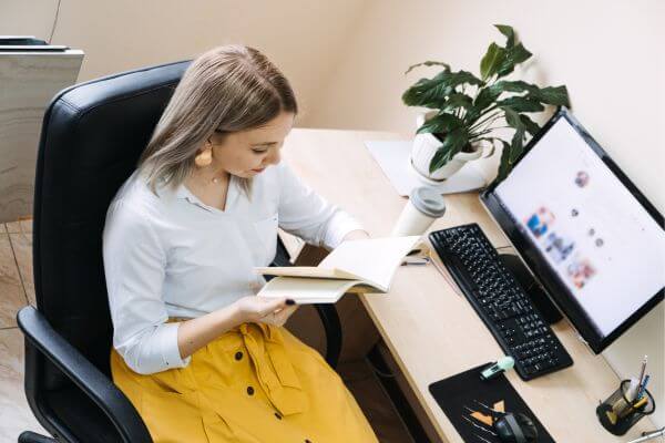 The picture of a woman reading a book together with a personal computer set up for work. 