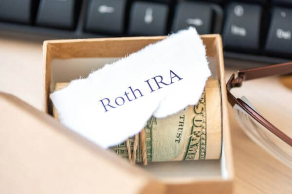 Image of a box filled with money and Roth IRA text and a eyeglass with keyboard around.